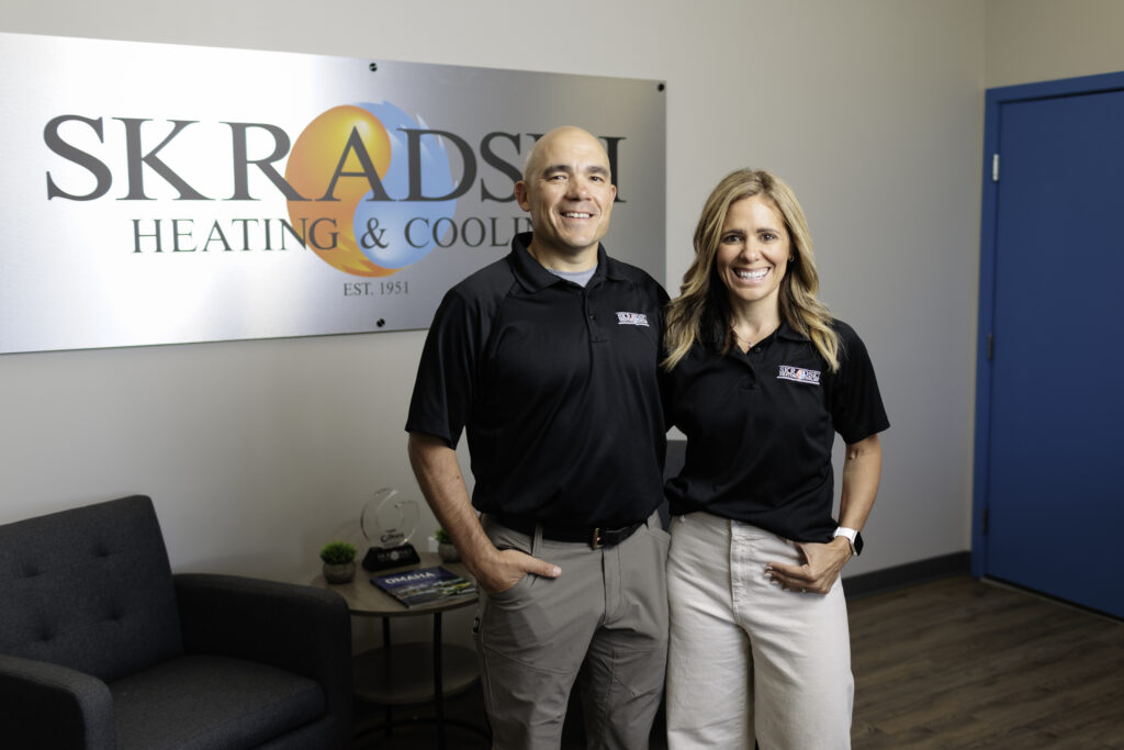Two people wearing black company shirts stand together and smile inside an office with a SKRADSKI Heating & Cooling sign on the wall behind them.