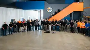 A large group of people pose for a photo inside a spacious industrial building, some standing on the ground and others on an orange staircase.