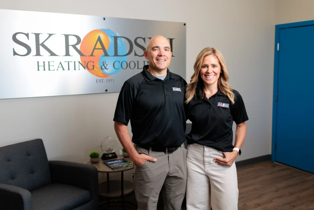 Two people in matching black polos stand smiling in an office next to a sign that reads "Skradski Heating & Cooling.