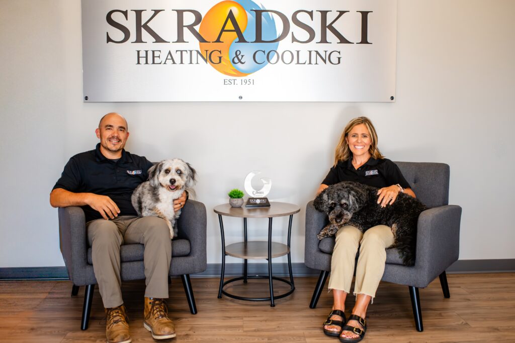 Two people sit in armchairs with two dogs in front of a "Skradski Heating & Cooling" sign. A small table with a trophy and plant is between them.