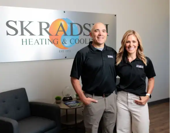 Two people stand smiling in front of a Skradski Heating & Cooling sign inside an office waiting area.