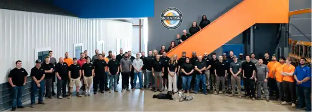 Group of people standing together in a large indoor space with the Skarboski logo on the wall and orange stairs in the background.