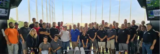 A large group of people pose together indoors at a driving range, standing and sitting in rows with golf nets visible in the background.